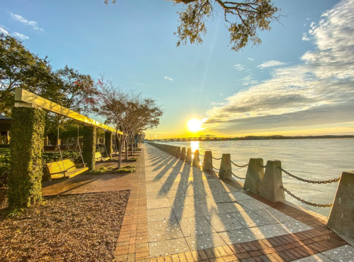 A serene waterfront scene at sunset, with trees, a walkway, and long shadows cast by the setting sun.