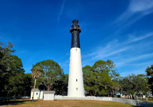 A tall lighthouse with a black and white design stands against a clear blue sky, surrounded by trees and grass.