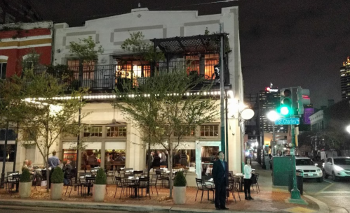 A lively restaurant at night, with outdoor seating and city lights in the background. People are gathered outside.