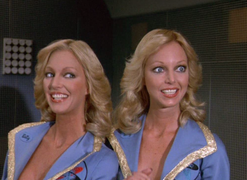 Two women with blonde hair and matching blue outfits smile in a retro setting, showcasing a playful and cheerful vibe.