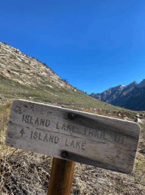 Wooden trail sign for Island Lake, indicating a distance of 2 miles, with mountains and blue sky in the background.