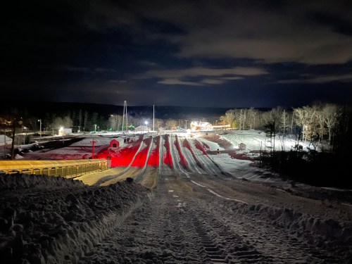 Nighttime view of a snow tubing hill, illuminated with lights, featuring snow-covered lanes and a cozy lodge in the background.