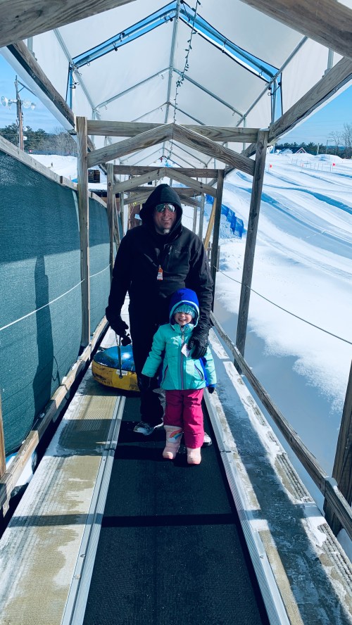 A man and a child stand on a snowy walkway, ready for tubing, both dressed warmly in winter gear.