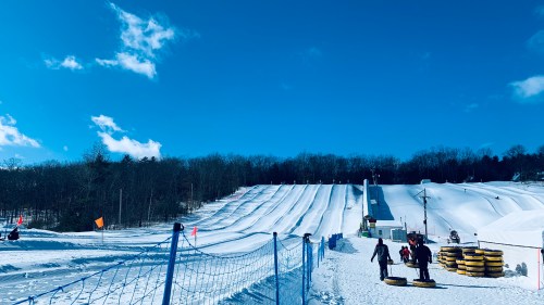 A snowy hill with multiple tubing lanes, people enjoying winter activities, and a clear blue sky in the background.