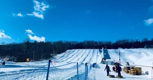 A snowy hill with multiple tubing lanes, people walking with tubes, and a clear blue sky in the background.