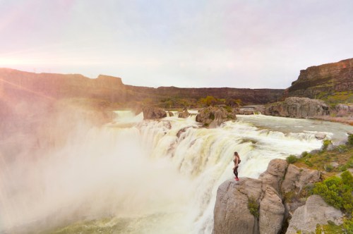 A person stands on a rocky ledge near a powerful waterfall, surrounded by lush greenery and dramatic cliffs.