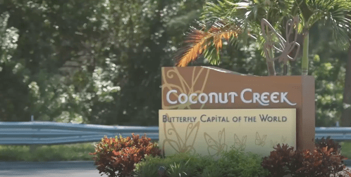 Sign for Coconut Creek, labeled "Butterfly Capital of the World," surrounded by lush greenery and colorful plants.