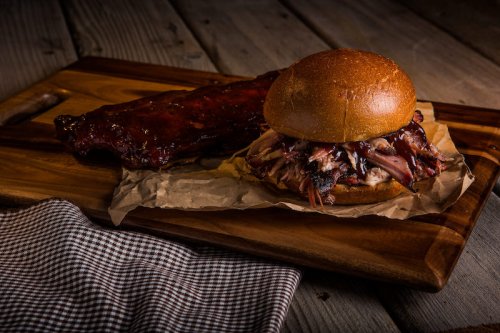 A pulled pork sandwich on a wooden board, accompanied by a side of glazed ribs and a checkered napkin.