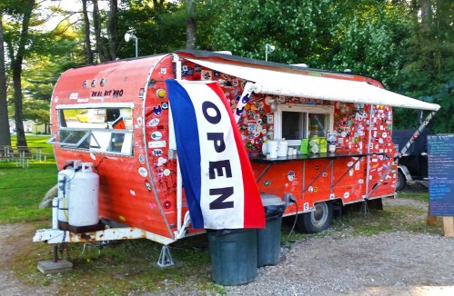 A vibrant red food truck with stickers, an "OPEN" sign, and a shaded serving window in a green outdoor setting.