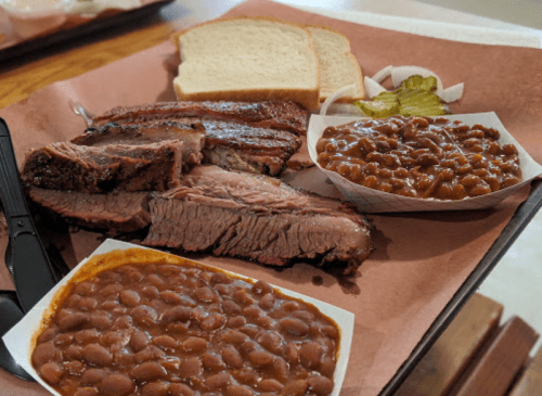 A plate of sliced brisket with bread, pickles, onions, and two servings of baked beans on a tray.