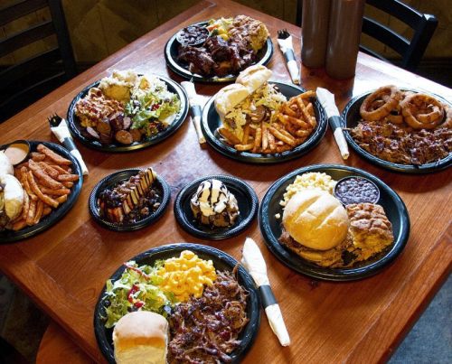 A table filled with various plates of barbecue dishes, sides, and desserts, including salads, sweet potatoes, and burgers.