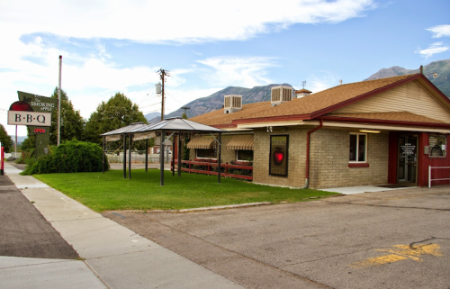 A BBQ restaurant with outdoor seating, surrounded by grass and mountains in the background, under a clear blue sky.