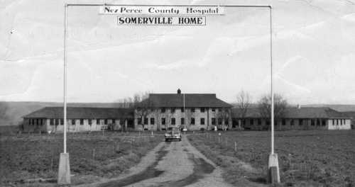 Black and white photo of Nez Perce County Hospital entrance, featuring a long driveway and a large building in the background.