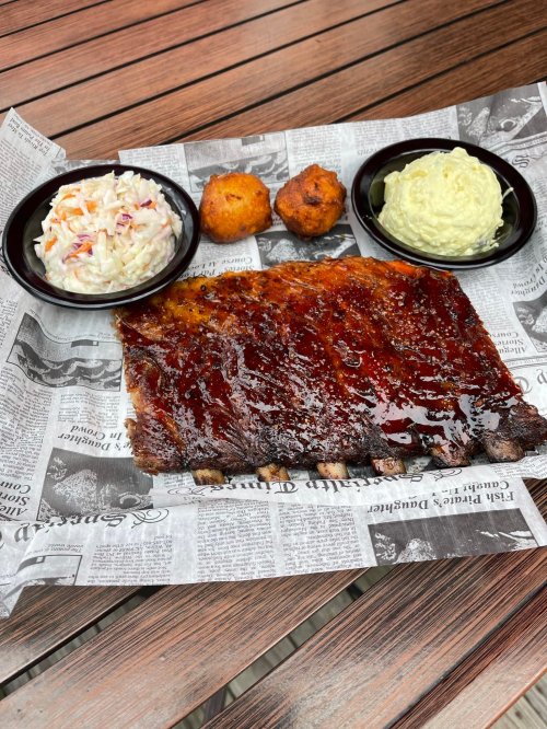 A plate of barbecue ribs with coleslaw, cornbread, and creamy dip, served on newspaper on a wooden table.