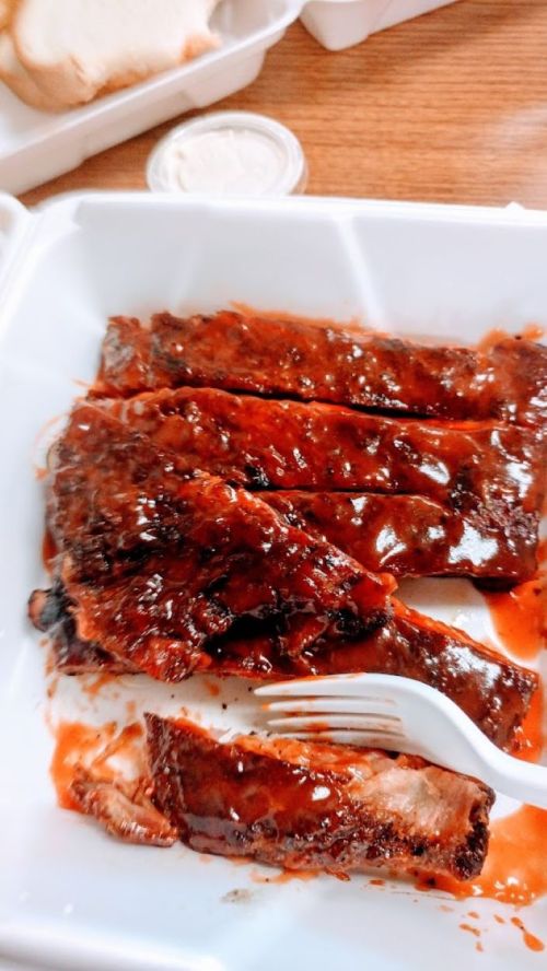 A takeout container with several pieces of barbecue ribs covered in sauce, accompanied by a plastic fork.