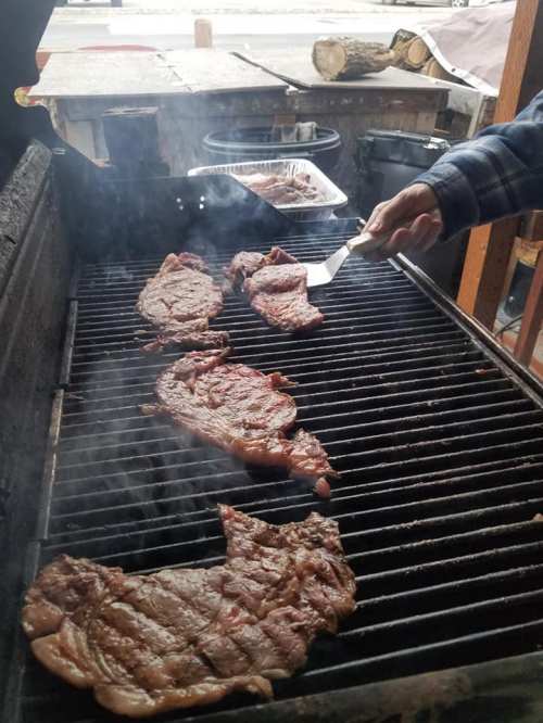 A hand using a spatula to flip steaks on a smoky grill outdoors.