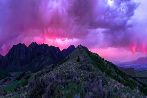 A panoramic view of mountains under a vibrant purple and pink sky, with lush green hills in the foreground.