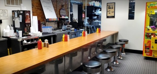 A diner interior featuring a long counter with stools, condiments, and a vintage arcade machine in the background.