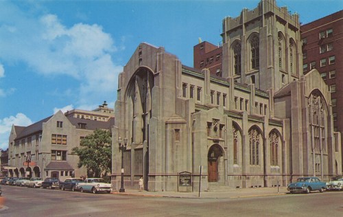 Historic stone church with Gothic architecture, surrounded by buildings and vintage cars on a sunny day.