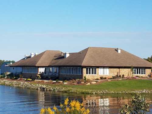 A brick building with a sloped roof beside a calm lake, surrounded by green grass and yellow flowers.