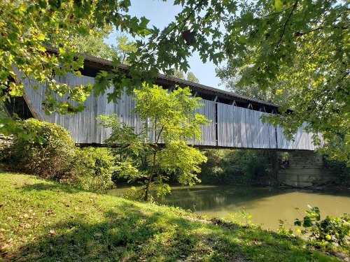 A covered bridge spans a calm river, surrounded by lush greenery and trees under a clear blue sky.
