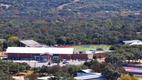 Aerial view of a sports complex surrounded by greenery, featuring a field, bleachers, and nearby buildings.