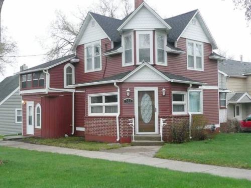 A two-story red house with white trim, featuring a front porch and a large bay window, set on a grassy lawn.
