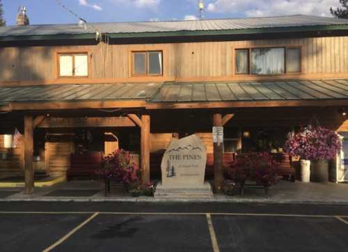 A rustic wooden building with a green metal roof, featuring flower baskets and a sign that reads "The Pines."