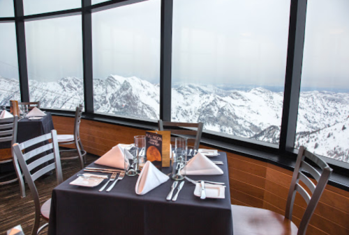 A dining table set with elegant tableware, overlooking snowy mountains through large windows.
