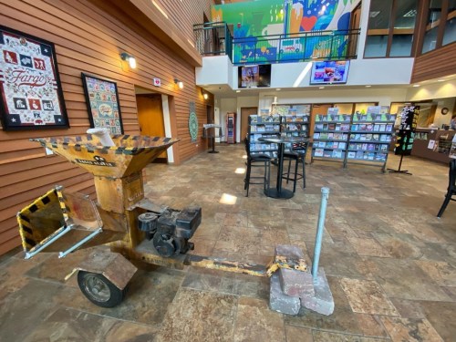 A rustic wood chipper on wheels in a spacious lobby with colorful artwork and shelves of books in the background.