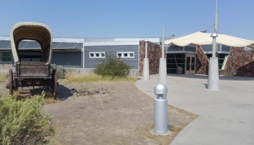 A historic wagon sits beside a modern building entrance with a shaded area and landscaping.