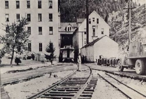 Black and white photo of a railway track with buildings and a vehicle in a mountainous area. A person stands on the tracks.