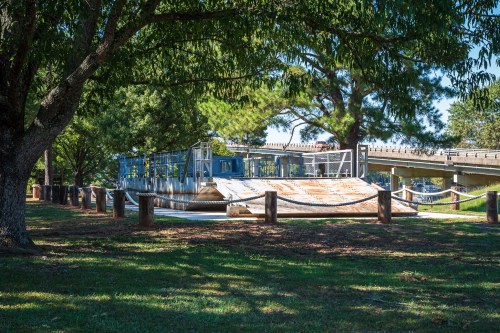 A view of a park with a wooden structure, trees, and a bridge in the background under clear blue skies.