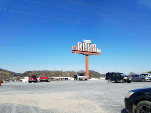 A large sign reading "Flea Market" stands above a gravel parking lot filled with cars under a clear blue sky.