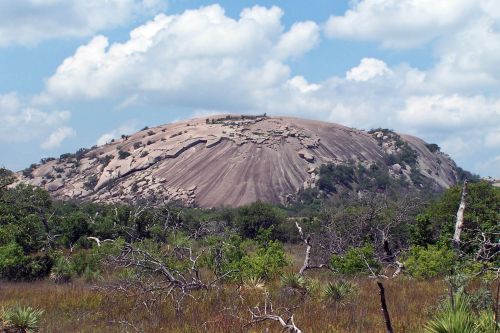A large, smooth rock formation rises above a grassy landscape under a partly cloudy sky. Sparse trees are in the foreground.