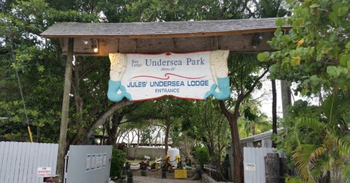 Sign for Jules' Undersea Lodge at Key Largo, surrounded by trees and tropical plants, marking the entrance to the park.