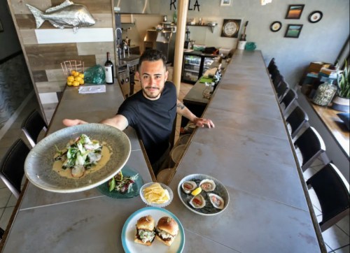 A chef presents a plate of food at a modern restaurant bar, with various dishes displayed on the counter.