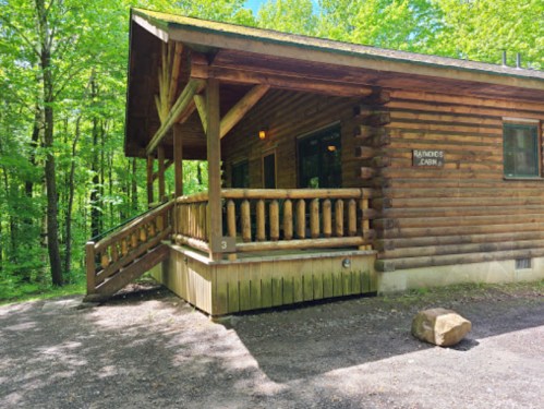 A wooden cabin with a porch, surrounded by lush green trees and a gravel path.