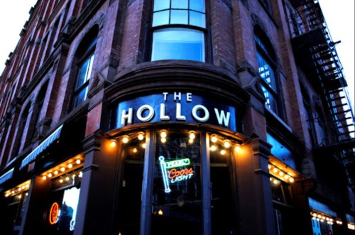 Exterior of a brick building with a sign reading "The Hollow" and neon lights, showcasing a lively atmosphere.
