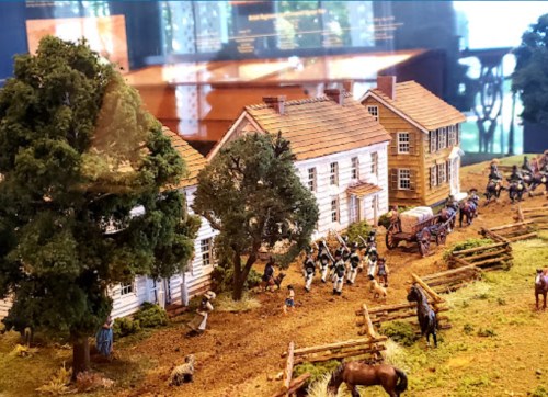 A detailed miniature scene depicting a historical settlement with buildings, trees, and figures engaged in various activities.