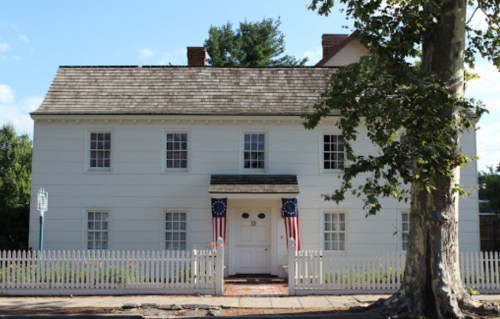 A white two-story house with a picket fence, featuring American flags and a large tree in front.