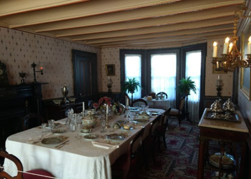 A vintage dining room with a large table set for a meal, surrounded by elegant decor and natural light from windows.