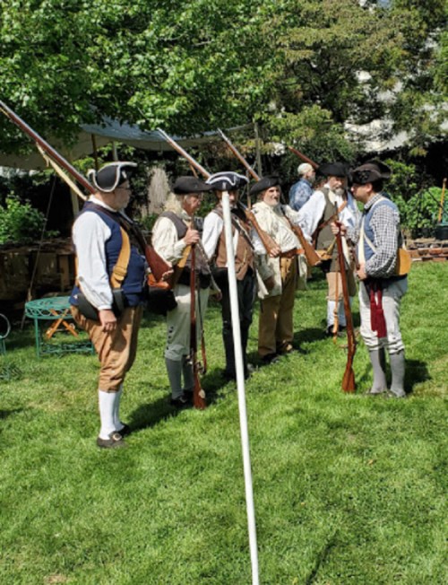A group of men in colonial attire, holding muskets, gather outdoors for a historical reenactment.