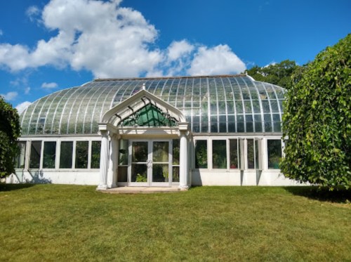 A large glass greenhouse with a white frame, surrounded by green grass and trees under a blue sky with clouds.