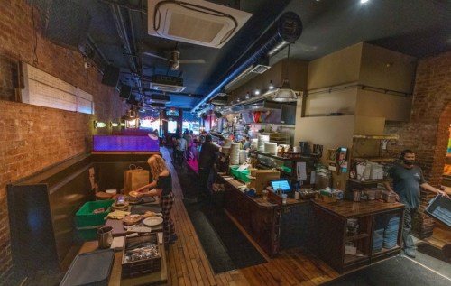 A bustling restaurant interior with exposed brick walls, a busy kitchen, and staff preparing food and serving customers.