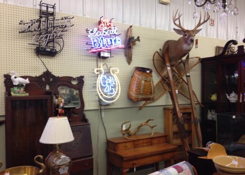 A collection of vintage items, including neon signs, a mounted deer head, and wooden furniture, displayed on a wall.