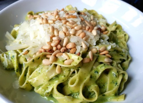 A bowl of fettuccine pasta topped with pine nuts and grated cheese, garnished with a green pesto sauce.