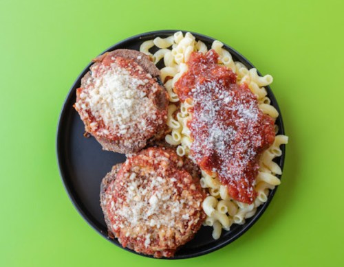 A black plate with two meatballs topped with marinara sauce and cheese, served with pasta and additional sauce.