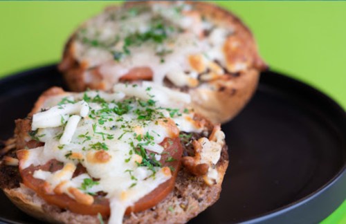 Two toasted halves of a bun topped with melted cheese, tomatoes, and herbs on a black plate against a green background.