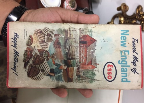 A hand holding a vintage travel map of New England, featuring illustrations of fishing and coastal scenes.